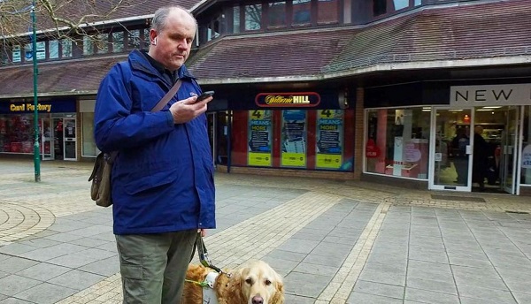 Persona ciega en la calle con un perro lazarillo y usando un smartphone.