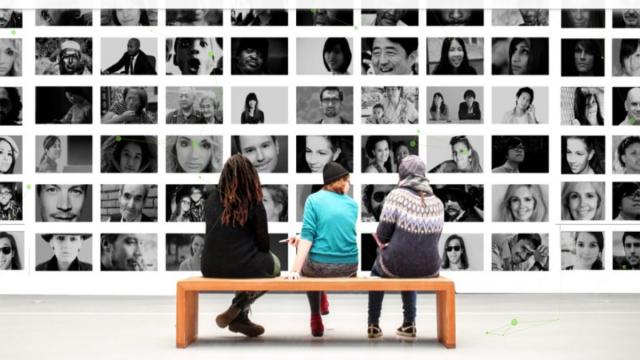 Tres mujeres de espaldas sentadas en un banco mirando fotografías en una pared.