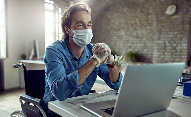 Un home en cadeira de rodas traballa no seu computador con máscara e luvas.